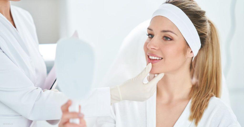 Nurse injector is touching a patient's jaw (MODELS) in the medical spa. The patient is looking at a mirror and enjoying her Masseter Dysport® treatment results.